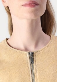 Veste jaune clair avec une texture lisse et semblable à des poils. Elle dispose d'une fermeture éclair argentée et d'un col rond, mettant en valeur de fines coutures.