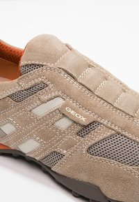 Beige mockaklädsel och mesh-sneakers med sydda detaljer, märkt med "GEOX"-etikett och orange inredning, vinklad för att visa sid- och toppsektioner.