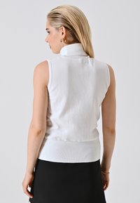 Vrouw met blond haar in een witte mouwloze coltrui en een zwarte rok, met haar rug naar de camera gekeerd tegen een effen achtergrond.
