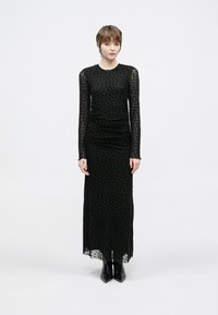 Melnas garām piedurknēm kleita ar polka punktu tīkliņa pārklājumu, ručētu vidukli un taisnu siluetu, kopā ar melnām potīšu zābakiem.