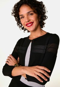 Zwarte cardigan met doorzichtige paneeldetails, aansluitend ontwerp, ronde halslijn en lange mouwen. Draag het over een grijze top, geaccessoireerd met een gouden horloge.