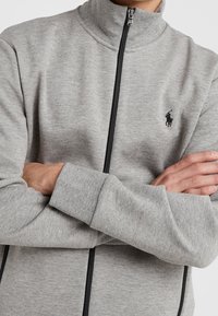 Světlé šedé zipové sako s vysokým límcem, obsahující černý zip, žebrované manžety a malé vyšívané logo na hrudi.