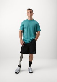 Camiseta de manga corta turquesa, pantalones cortos negros, zapatillas deportivas grises. Presenta una pierna prostética con acabado metálico. Fondo neutral.