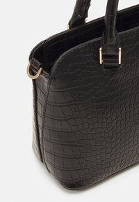 Sac à main noir à texture crocodile, avec anneaux en métal doré et deux poignées courtes sur fond blanc.