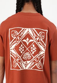Tricou din bumbac de culoare ruginie, prezentând un design grafic mare de culoare albă pe spate, care ilustrează un copac și motive florale într-o formă de diamant.