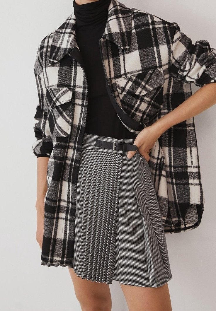 Veste oversize à carreaux noirs et blancs portée sur un col roulé noir, associée à une mini-jupe plissée noire et blanche avec une ceinture.