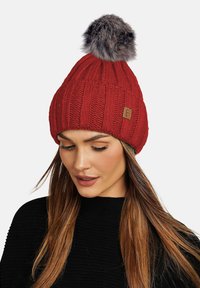 Red knitted beanie with vertical ribbing, featuring a fluffy gray pom-pom on top and a small logo patch on the side.