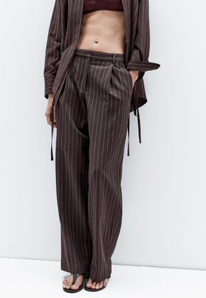 Braune Nadelstreifen-Hosen mit entspannter Passform, ausgestattet mit Falten und hoher Taille. Kombiniert mit einem dunklen Oberteil und schlichten Sandalen.