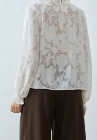 Blouse blanche transparente avec des motifs floraux, poignets froncés et col haut. Portée sur un pantalon marron foncé, mettant en avant un design léger et aérien.