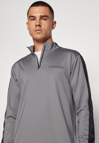 Grau athletischer Pullover mit einem Viertel-Reißverschluss, der einen leichten Glanz, schwarze seitliche Akzente und ein Hummel-Logo auf der Brust aufweist.