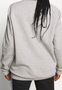 Persona con cabello largo trenzado vista desde atrás, lleva un sudadera gris claro y pantalones negros con franjas blancas a los lados.