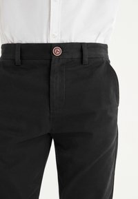 Pantalon noir taille haute avec un bouton marron et des passants pour ceinture, porté avec une chemise blanche rentrée.