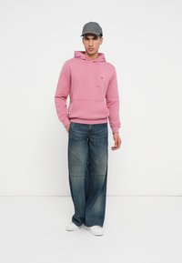 Jasnoróżowa bluza z kapturem z przednią kieszenią, zestawiona z luźnymi, wyblakłymi niebieskimi dżinsami. Model nosi szaro-czapkę i białe trampki.