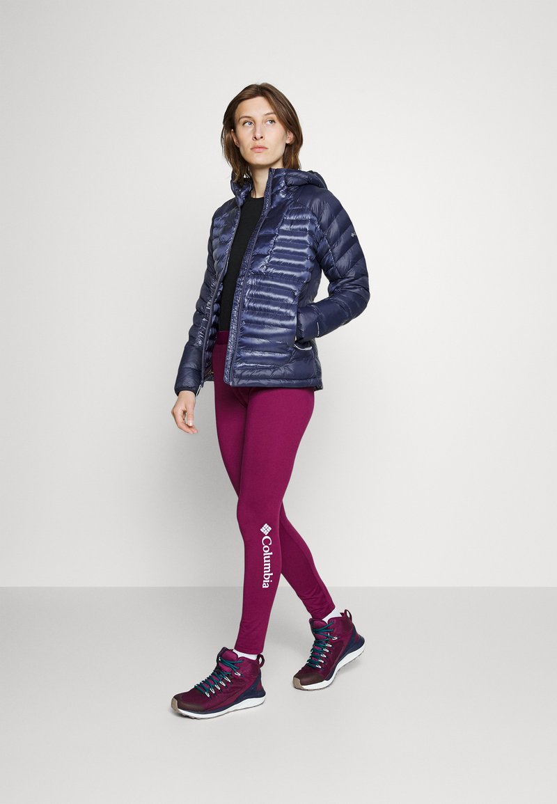 Doudoune marine, design ajusté, avec capuche ; associée à un legging bordeaux avec logo ; chaussures montantes marron et marine avec lacets.