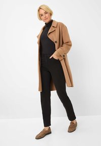 Cappotto in lana color marrone chiaro con grandi bottoni, dolcevita nero, jeans neri e scarpe con stampa leopardo. La modella è in posa con una mano in tasca.