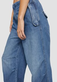 Blauwe denim broek met een losse fit, voorzien van zijzakken en een licht vervaagde textuur. Stikseldetails zijn zichtbaar langs de naden.