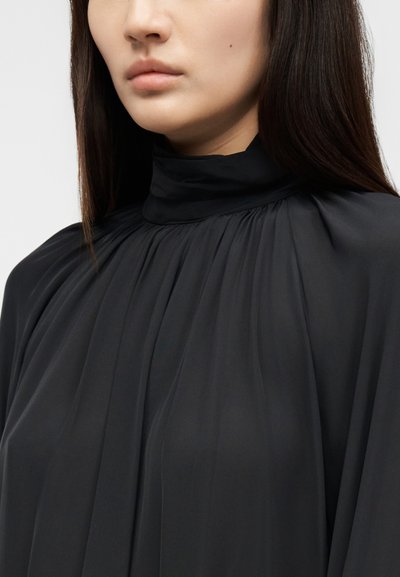 Femme portant un chemisier noir à col montant et manches longues avec des plis de tissu rassemblé autour du col et de la poitrine.