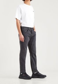 Jeans de mezclilla negros, ajuste recto, usados con una camiseta blanca. Las zapatillas negras presentan un branding sutil. Diseño limpio sin patrones.