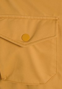 Senapsfärgad tyg med texturerad yta, med en klaffficka som säkras av en rund, matchande knapp. Enkel och praktisk design.