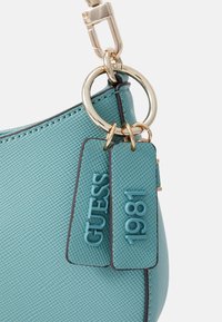 Tyrkysová strukturovaná kabelka s kroužkem v zlaté barvě a dvěma visačkami s ražbou "GUESS" a "1981".