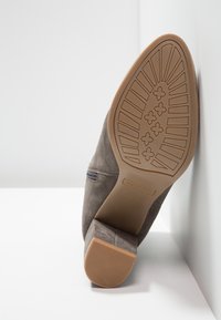 Bottine en daim marron inclinée contre un mur blanc, montrant la semelle avec des motifs géométriques et en croix ainsi que le talon carré.