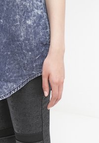 Top in denim blu sbagliato con orlo irregolare, abbinato a leggings neri testurizzati con dettagli neri in accentuazione. Mano appoggiata sui leggings.