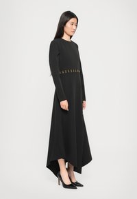 Black long-sleeve dress with a high-low hem, featuring a gold accent belt. The material appears smooth and structured. Worn with black heels.