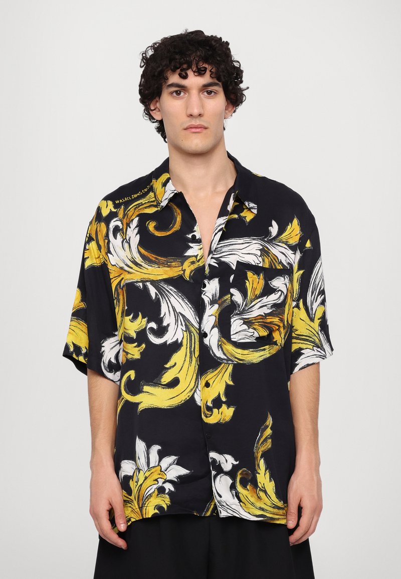 Korte mouwen shirt van zwarte stof met grote gele en witte bloemenpatronen, een knoopsluiting aan de voorkant en een borstzak.