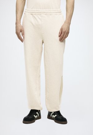 Mand iført lyse beige joggingbukser med elastisk talje og sorte sneakers står mod hvid baggrund.