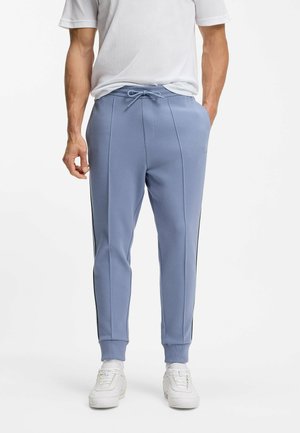 Mand iført lyseblå taperede joggingbukser med sidelommer, manchetter ved anklerne, hvide sneakers og hvid kortærmet skjorte, hænderne i lommerne.