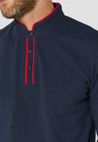 Marinblå polo tshirt med mandarin-krage, kompletterad med röda detaljer och en treknappsslutning. Texturerad tyg med en slät finish.