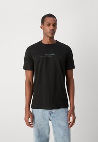 Juodas medvilnės marškinėliai trumpomis rankovėmis ir apvaliu iškirpimu. Priekinėje pusėje yra „Calvin Klein Jeans“ logotipas baltos ir šviesiai mėlynos spalvos tekstu.