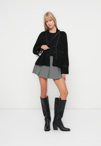 Cardigan noir oversize avec des clous, mini jupe plissée grise, et bottes noires en cuir à talons blocs jusqu'aux genoux. Petit sac à main inclus.