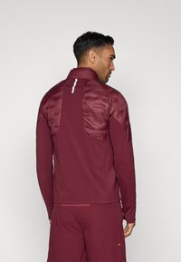 Bordový športový kabátik s vypchávkou na chrbte, vysokým golierom, dlhými rukávmi a reflexným prúžkom. V páre s dokonale zladenými bordovými nohavicami.