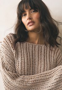 Lysebeige strikket sweater med løst, åbent vævemønster, der har brede ærmer og en afslappet halsudskæring. Blødt, tekstureret stof.