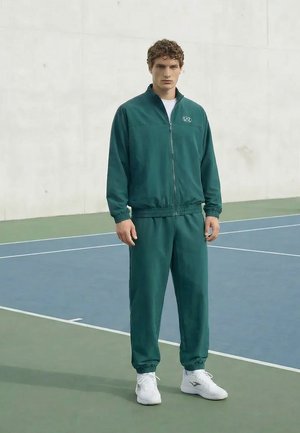 Jeune homme debout sur un court de tennis, portant un survêtement vert foncé et des baskets blanches, faisant face à l'avant avec une expression neutre.