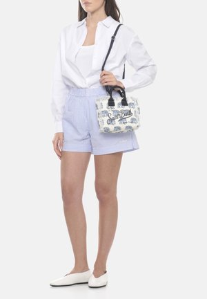 Donna che indossa una camicia bianca con bottoni, pantaloncini a righe azzurro chiaro, ballerine bianche, che tiene una piccola borsa bianca e blu con motivo sulla spalla.