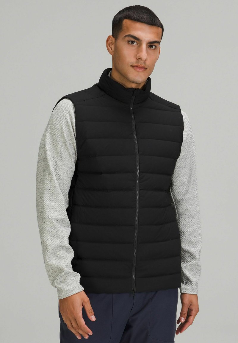 Schwarze Weste aus Pufferstoff mit hohem Kragen und Reißverschluss über einem strukturierten, hellgrauen Langarmshirt, das elastische Bündchen und eine lockere Passform hat.