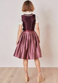 Femme debout de dos, portant un gilet violet avec des manches en dentelle blanche et une jupe midi brillante à motifs, avec des sandales compensées sur un sol en bois.