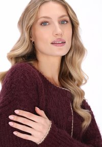 Kvinde med bølget blond hår iført en dyb lilla fuzzy knapstrikket sweater, der krydser armene med lys rosa manicure.