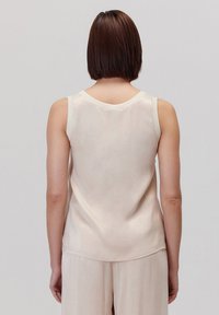 Bez rękawów, jasnobeżowy top na ramiączkach wykonany z miękkiego materiału. Posiada okrągły dekolt i gładki, minimalistyczny fason z lekko zaokrąglonym dołem.