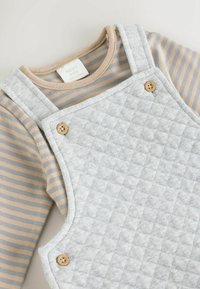 Grau gesteppte Latzhose mit Holzknöpfen über einem beige-grauen gestreiften Langarmshirt. Weiches Baumwollmaterial. Etikett sichtbar.