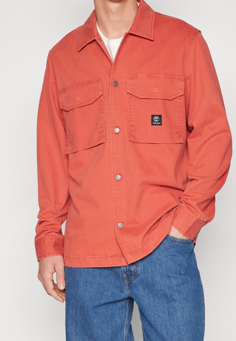 Röd-orange canvas skjorta med två bröstfickor, tryckknappar och en liten svart logobläck på vänstra fickan. Buren över blå jeans.