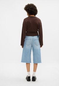 Suéter castanho brilhante de mangas longas combinado com calções de ganga de perna larga de tom azul claro. Sapatos pretos e meias brancas com padrão visíveis.