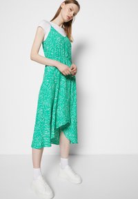 Vestido verde com um padrão branco ondulado, apresentando um corte solto e uma bainha assimétrica. Combinado com ténis brancos e uma camisa branca por baixo.