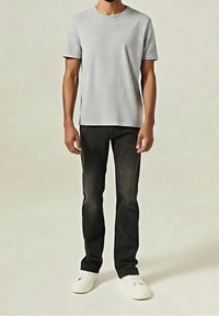 Homme portant un t-shirt gris clair à manches courtes, un jean noir et des baskets blanches, debout devant un fond clair uni.