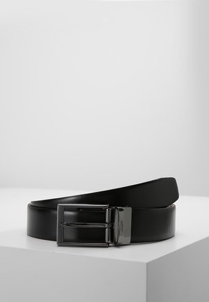 Ceinture en cuir noir avec une boucle rectangulaire en argent, roulée et posée sur une surface blanche sur fond gris uni.