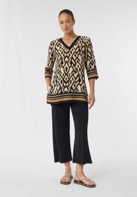 Musteroberteil mit V-Ausschnitt, dreiviertel Ärmeln, das ein geometrisches Design in Schwarz, Creme und Braun aufweist. Kombiniert mit langen schwarzen Hosen und Riemchensandalen.