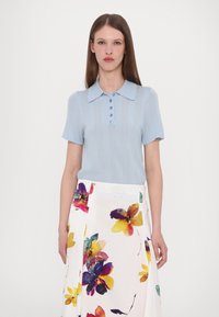 Lichtblauwe polo met korte mouwen met ribbelstructuur en knoopsluiting, gecombineerd met een witte rok met een kleurrijk bloemenpatroon.