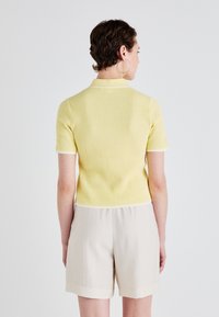 Polo a maniche corte, a coste, giallo con rifiniture bianche, abbinato a pantaloni corti beige. La texture è liscia, con una forma aderente.